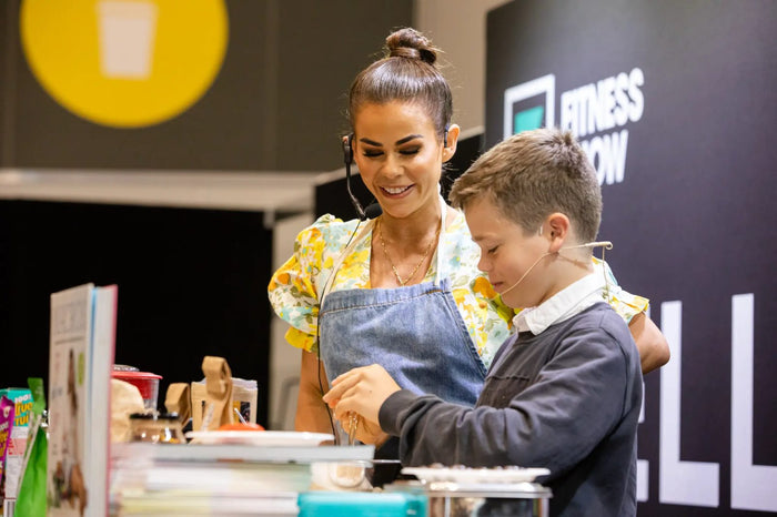 Sophie Guidolin demonstrating healthy cooking with son at the Melbourne Fitness Show, wearing a denim apron and headset microphone.